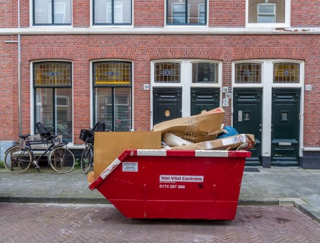 A large red skip bin, with a white label displaying contact information, is positioned on a paved sidewalk in front of a row of brick terraced houses with dark green doors in Woolwich. The bin is filled with various discarded items including flattened cardboard boxes, plastic bags, and other general waste materials, some of which are spilling over the top. To the left of the bin, there are two bicycles parked against the building's wall, one with a black frame and the other with a grey frame, both secured with locks. The background features the building’s brick facade with evenly spaced windows that have white frames and yellow security bars, reflecting a typical residential street scene. The environment appears to be in daylight, with natural light illuminating the scene, emphasizing the textures of the brickwork, metal bin, and waste materials. This image relates to commercial waste removal services provided by [COMPANY_NAME], serving Woolwich and the surrounding areas with rubbish collection and clearance solutions.