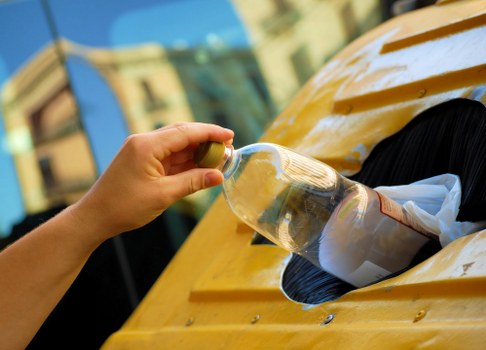 A person’s hand holding a clear plastic bottle filled with a dark liquid over a large yellow recycling or waste disposal bin, which has a circular opening with a black liner or bag inside. The bin is situated on a paved exterior surface, possibly a driveway or sidewalk, with a blurred background of buildings and blue sky. The scene appears to be part of a waste collection or rubbish removal process, with the focus on the act of discarding the bottle as part of general waste or recycling, relevant to services such as those provided by Commercial Waste Removal Woolwich in the Woolwich area. The lighting suggests daytime with natural sunlight illuminating the scene, emphasizing the textures of the plastic bottle, the yellow painted surface of the bin, and the surrounding environment.