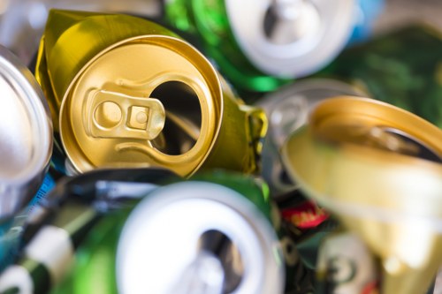 Close-up photograph of several aluminium beverage cans with open tops, positioned on top of each other. The cans display various colours including yellow, green, and silver, with some featuring pull tabs and others with open rings. The surface of the cans is slightly reflective, showing subtle highlights and shadows due to ambient lighting. The cans are situated outdoors on a paved or concrete surface, with a blurred background suggesting a recycling or rubbish disposal context. The image emphasizes the metallic textures and the condition of the cans, which may be relevant for waste collection or recycling services offered by Commercial Waste Removal Woolwich, serving areas near Woolwich in London postcodes.
