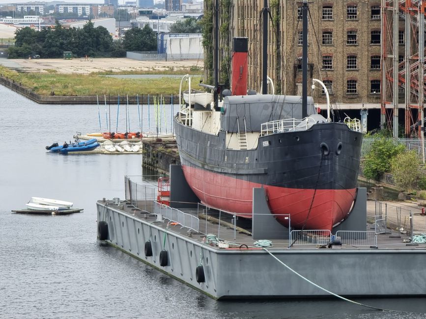 A large vintage-style boat with a black upper hull and a red lower hull is positioned above water, held in place on a floating concrete platform by metal supports and ropes. The boat features a white